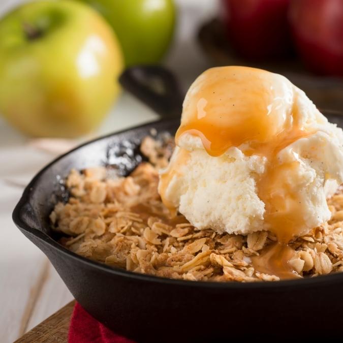 A close-up of warm apple crisp in a small dark dish, topped with a scoop of melting vanilla ice cream and a drizzle of Gravenstein Apple Balsamic.