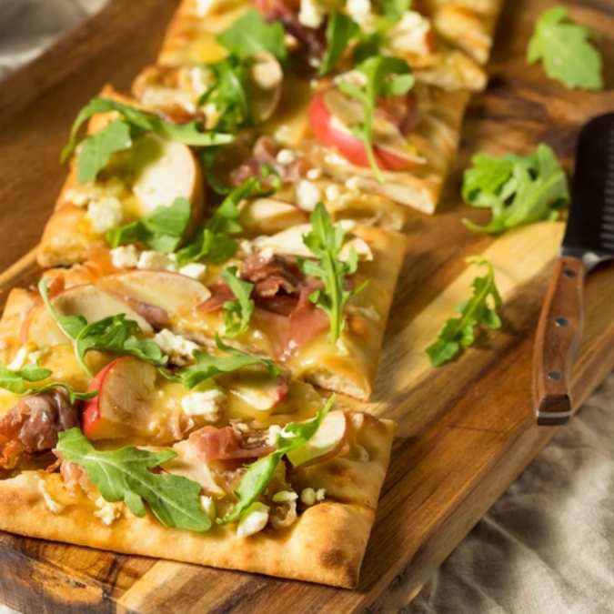 A sliced rectangular flatbread pizza topped with melted cheese, prosciutto, and fresh arugula.