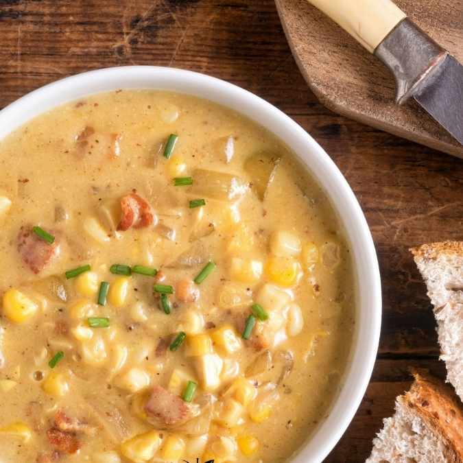 A white bowl filled with creamy corn chowder, garnished with bacon bits and fresh chives, sitting on a rustic wooden surface next to a slice of crusty bread, a wooden spoon, and a knife.
