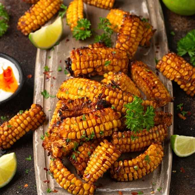 Golden, crispy roasted corn ribs seasoned with spices, served on a parchment-lined wooden board and garnished with fresh herbs and lime wedges.