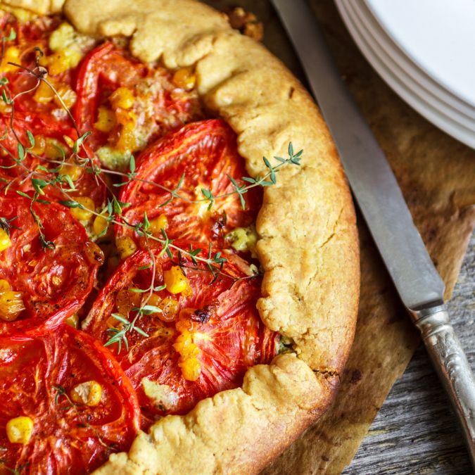 A rustic baked galette filled with sliced red tomatoes, sweet yellow corn, and fresh thyme sprigs, resting on parchment paper on a wooden surface next to white plates.