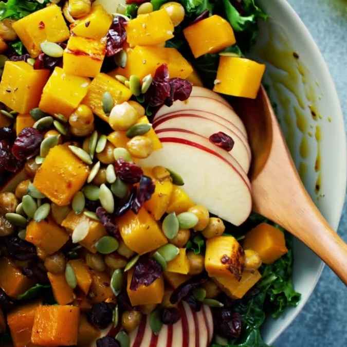 A vibrant fall harvest salad in a bowl, featuring a base of hearty green kale topped with golden roasted butternut squash, crisp apple slices, chickpeas, dried cranberries, crunchy pepitas, and creamy crumbled goat cheese.