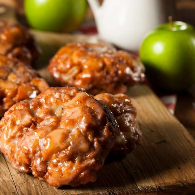 A close-up of golden brown, freshly glazed homemade apple fritters resting on a wooden surface, with bright green apples blurred in the background.