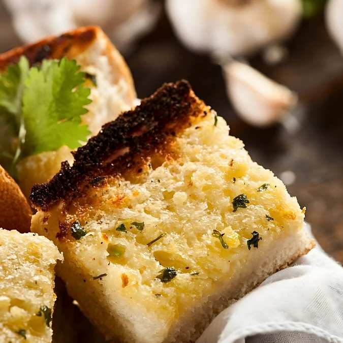 A close-up of a thick slice of golden-brown toasted garlic bread drizzled with olive oil and sprinkled with herbs.
