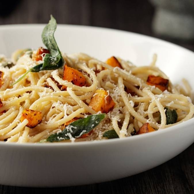 A white bowl filled with spaghetti tossed with roasted butternut squash cubes, sage leaves, and grated Parmesan cheese, resting on a dark wooden surface.