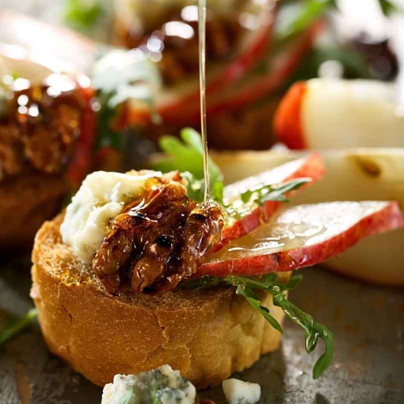 A piece of walnut and apple bruschetta with creamy gorgonzola on toasted bread, being drizzled with honey and balsamic.
