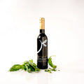 A bottle of red wine from 'The Little Shop' with a gold foil cap and a black label featuring a white stylized leaf logo, styled with fresh green basil leaves on a clean white background.