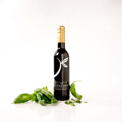 A bottle of red wine from 'The Little Shop' with a gold foil cap and a black label featuring a white stylized leaf logo, styled with fresh green basil leaves on a clean white background.