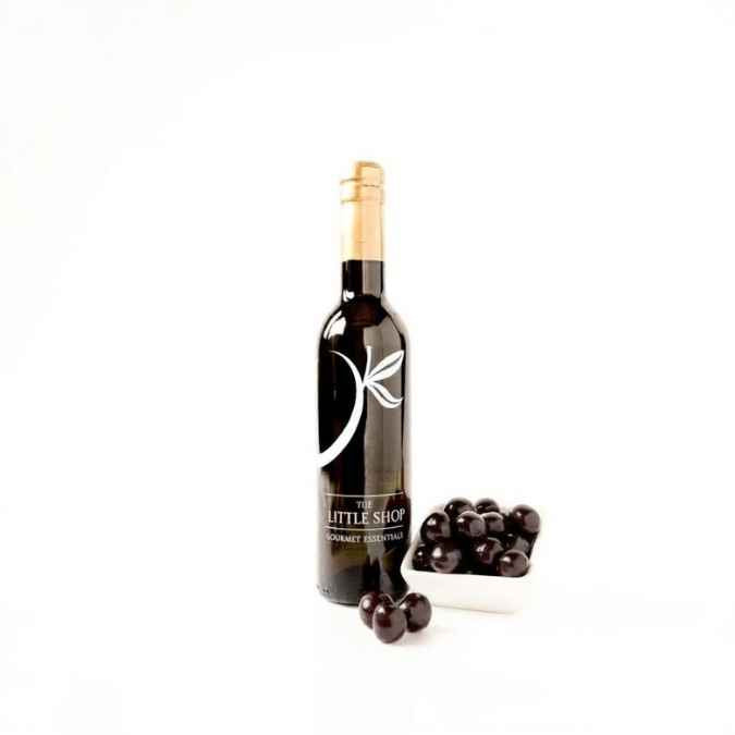 A dark glass bottle of balsamic vinegar with a gold foil top and a white leaf logo on the label, standing next to a small white dish of black cherries on a clean white background.