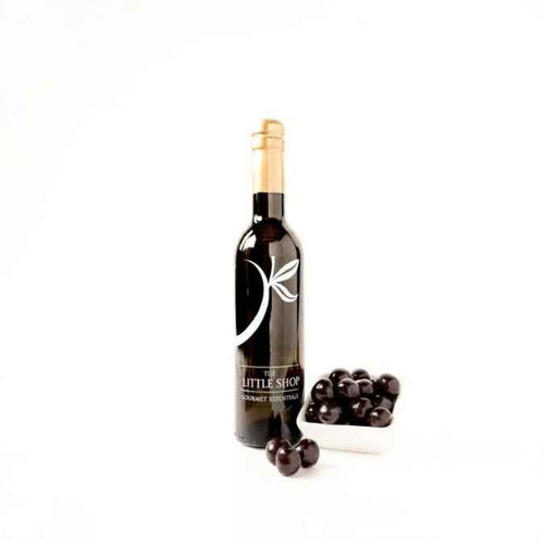 A dark glass bottle of balsamic vinegar with a gold foil top and a white leaf logo on the label, standing next to a small white dish of black cherries on a clean white background.