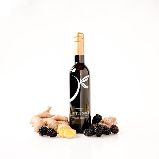 A bottle of gourmet balsamic vinegar by 'The Little Shop' is displayed on a white background, styled with fresh ingredients including ginger roots, sliced ginger, and blackberries.
