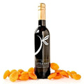 A dark glass bottle of 'The Little Shop Gourmet Essentials' with a gold foil neck wrap and white logo, surrounded by scattered dried orange apricots on a clean white background.