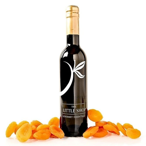 A dark glass bottle of 'The Little Shop Gourmet Essentials' with a gold foil neck wrap and white logo, surrounded by scattered dried orange apricots on a clean white background.