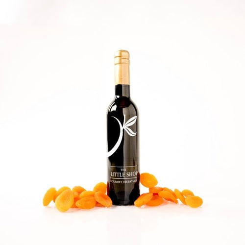 A dark glass bottle of gourmet balsamic vinegar on a reflective white surface with scattered apricot pieces styled around the bottle. 