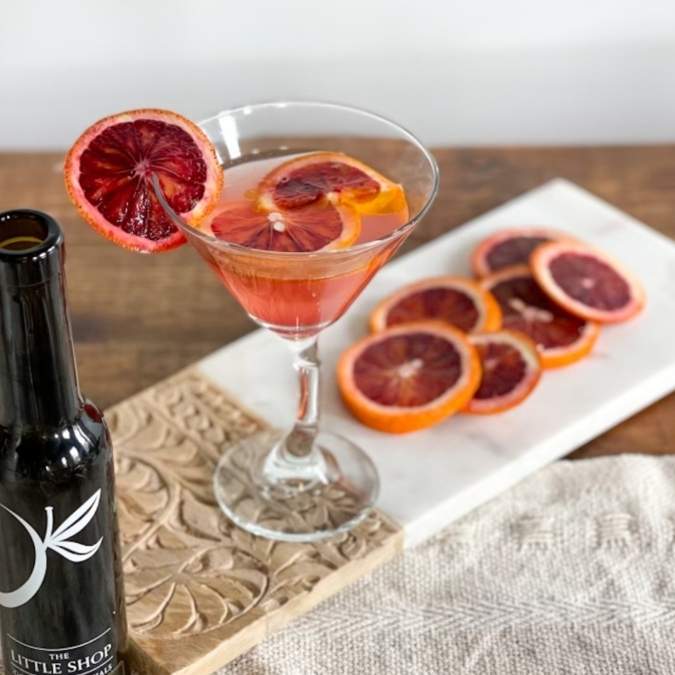 A pink cocktail in a martini glass garnished with sliced blood orange, set on a wooden table beside a dark bottle labeled 'The Little Shop' and a white platter with more orange slices.