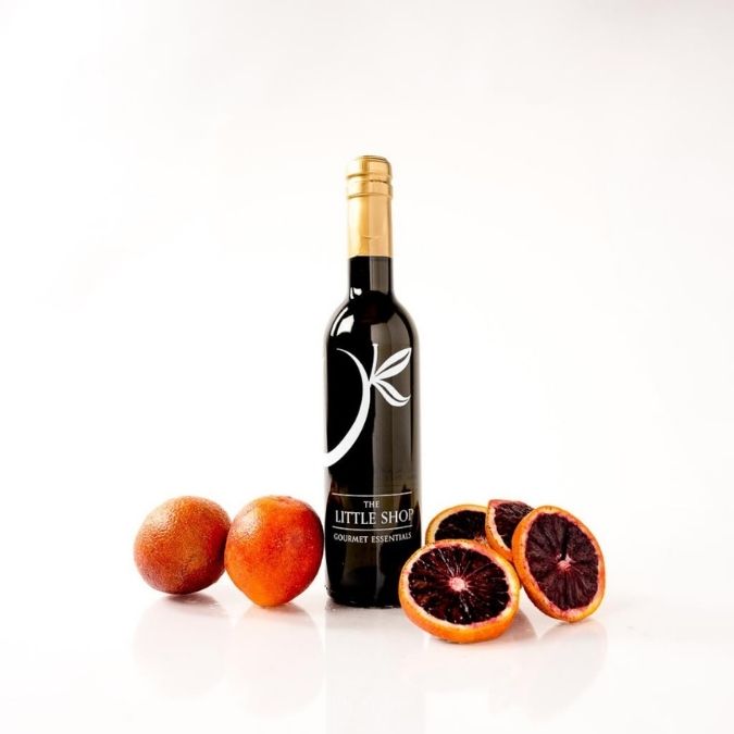 A bottle blood orange olive oil in a black bottle and gold cap stands on a white reflective surface, surrounded by whole and sliced blood oranges. The label reads 'THE LITTLE SHOP'.