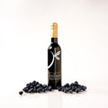 A bottle of balsamic vinegar centered on a white background, surrounded by scattered blueberries at the base. The bottle has a black label with a white stylized leaf logo.