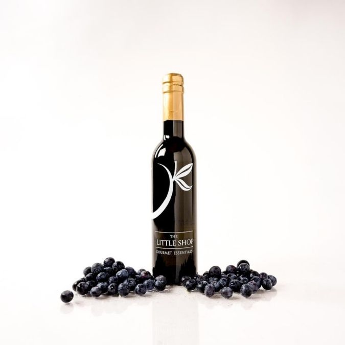 A bottle of balsamic vinegar centered on a white background, surrounded by scattered blueberries at the base. The bottle has a black label with a white stylized leaf logo.