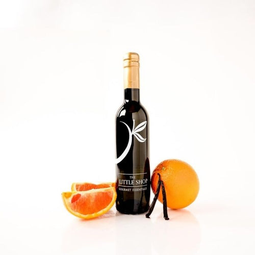 A dark glass bottle of gourmet balsamic vinegar on a white reflective surface with sliced oranges, vanilla beans, and a whole orange styled around the bottle. 