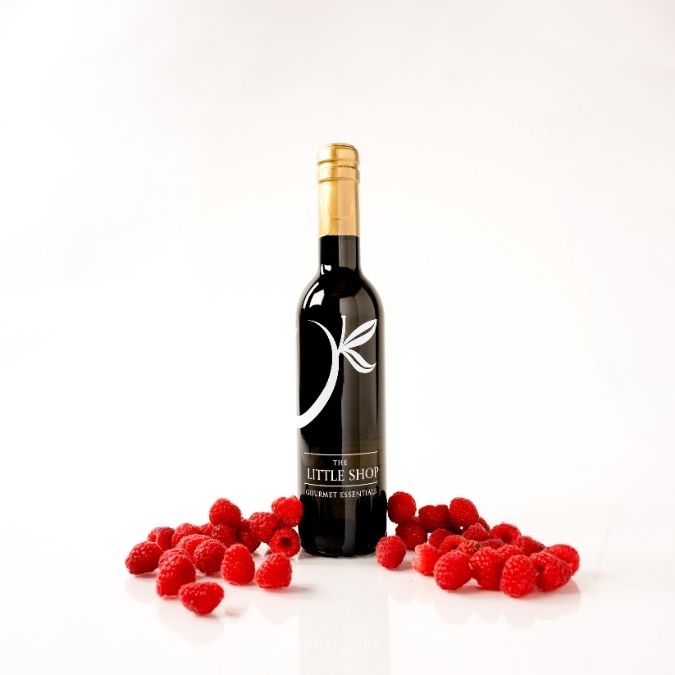 A dark glass bottle of balsamic vinegar with a gold foil top and a black label reading 'THE LITTLE SHOP', centered on a white background with scattered fresh red raspberries in front.