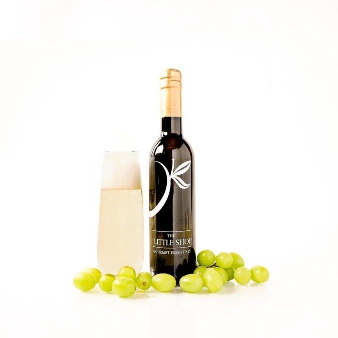 A bottle of gourmet white wine vinegar with a gold foil cap, standing next to a filled champagne flute, with green grapes arranged at the base on a white background.