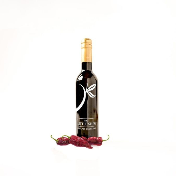 A dark glass bottle of olive oil with a gold foil top and a black label featuring a white leaf logo, standing on a white background with three red chipotle peppers at its base.
