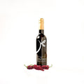 A dark glass bottle of olive oil with a gold foil top and a black label featuring a white leaf logo, standing on a white background with three red chipotle peppers at its base.