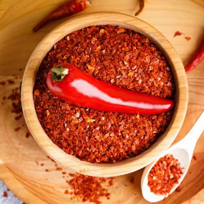 A wooden bowl filled with coarse chipotle pepper flakes, topped with a whole chipotle pepper, on a wooden surface with a small white spoon holding pepper flakes.