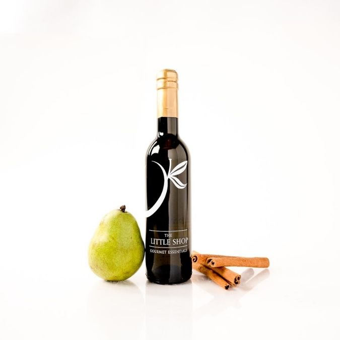 A bottle of The Little Shop gourmet balsamic vinegar next to a green pear and two cinnamon sticks on a white background.