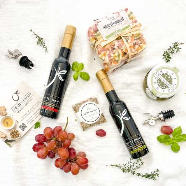 A styled flat-lay of a gourmet gift basket on a white textured fabric background, featuring two bottles of olive oil, a jar of honey, a bag of colorful pasta, a small soap bar, a card from 'The Little Shop', and fresh grapes and herbs.