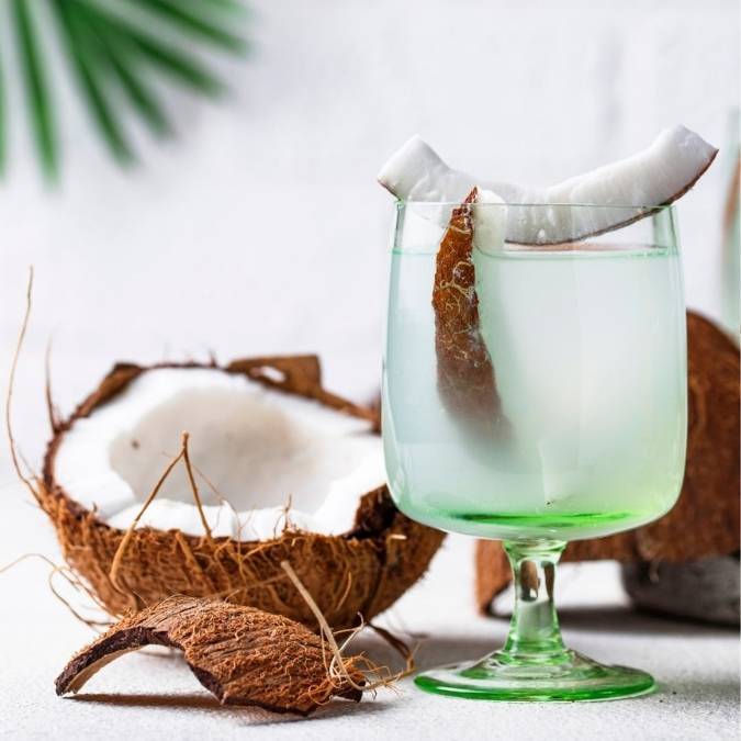 A clear stemmed glass with a green-tinted base filled with coconut water, garnished with coconut pieces, set beside halved coconuts on a light surface.