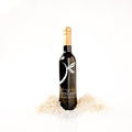 A dark glass bottle of balsamic vinegar with a gold foil top and a white stylized logo, standing upright in shredded coconut on a clean white background.