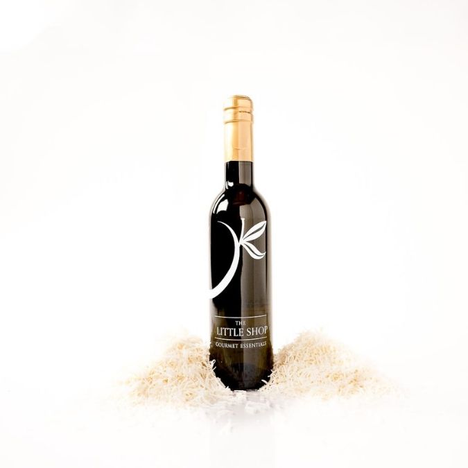 A dark glass bottle of balsamic vinegar with a gold foil top and a white stylized logo, standing upright in shredded coconut on a clean white background.