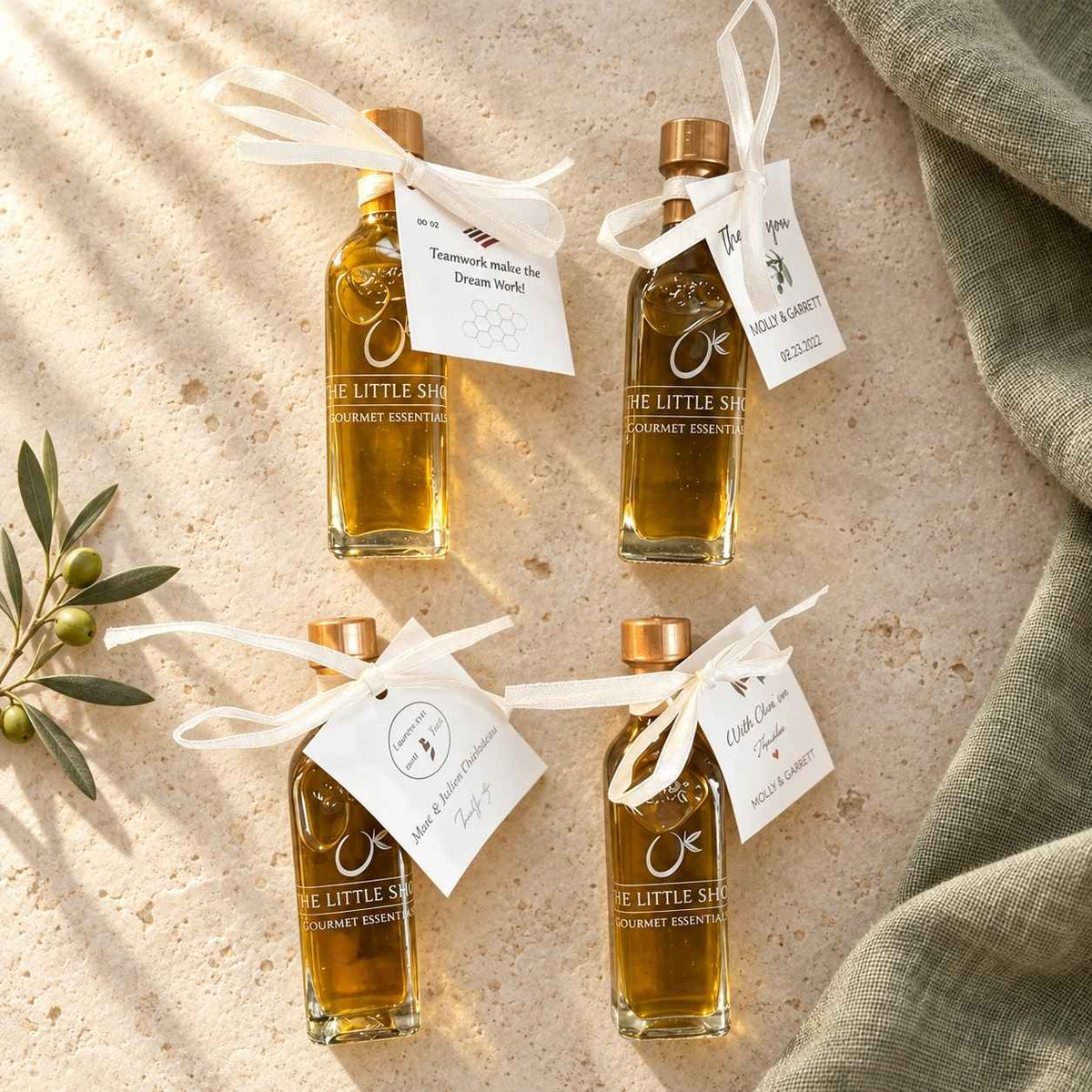 Four small glass bottles of corporate and event gift olive oil from The Little Shop of Olive Oils, tied with white satin ribbons and custom personalized gift tags, arranged on a neutral fabric background with an olive branch.