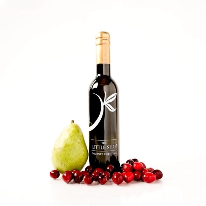 A dark glass bottle of wine with a gold foil top and a white leaf logo, styled with a green pear and red cranberries on a white background. The label reads 'THE LITTLE SHOP GOURMET ESSENTIALS'.