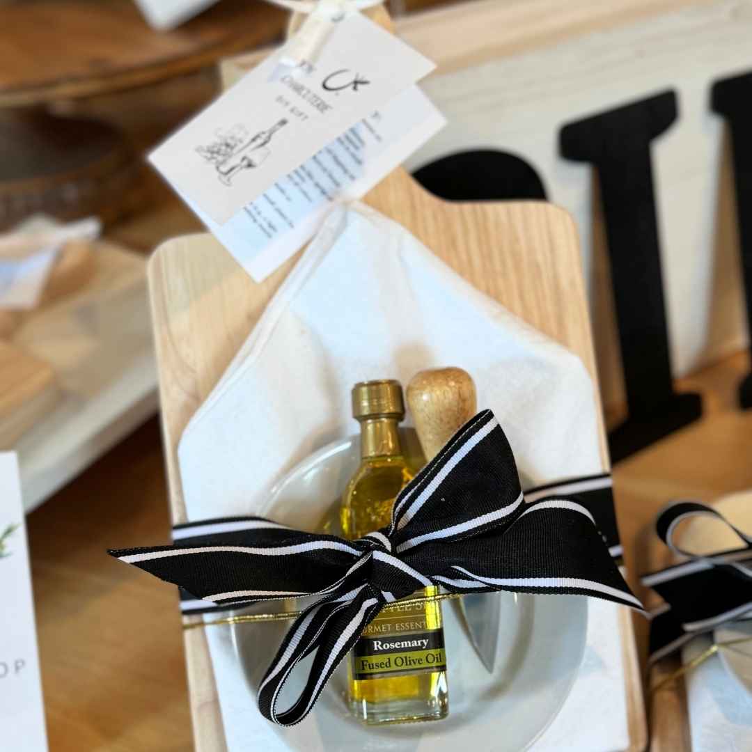 A small bottle of Rosemary Herb Balsam olive oil presented in a gift box with a white napkin and a black and white striped ribbon bow, on a wooden board.