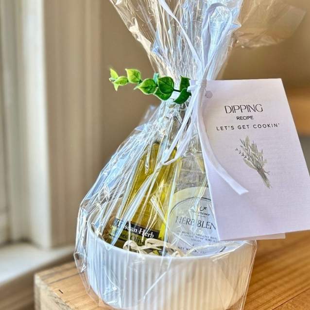 A gift dish filled with a bottle of olive oil, a bottle of wine, and a white ribbed bowl, wrapped in clear cellophane with a green sprig and a card that reads 'DIPPING RECIPE LET'S GET COOKIN''.