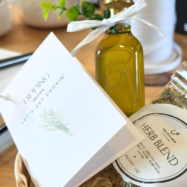 A styled gift set featuring a white recipe card that reads 'DIPPING recipe LETS GET COOKIN'', a glass bottle of olive oil tied with a white ribbon, and a jar of Thyme Shop Herb Blend in a wicker basket.