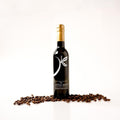 A bottle of The Little Shop balsamic vinegar with a gold foil cap and a black label, styled on a white reflective surface with scattered coffee beans in front of it.