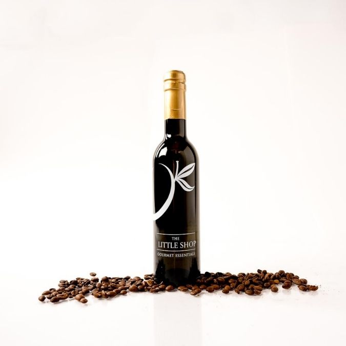 A bottle of The Little Shop balsamic vinegar with a gold foil cap and a black label, styled on a white reflective surface with scattered coffee beans in front of it.
