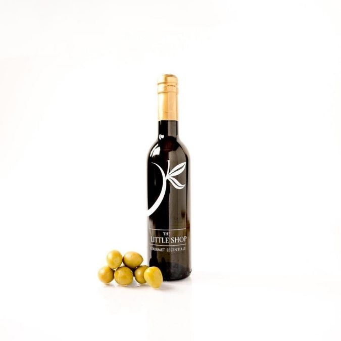 A dark glass bottle of extra virgin olive oil with a gold foil top and a white leaf logo, standing on a white background next to several green olives. The label reads 'THE LITTLE SHOP'.