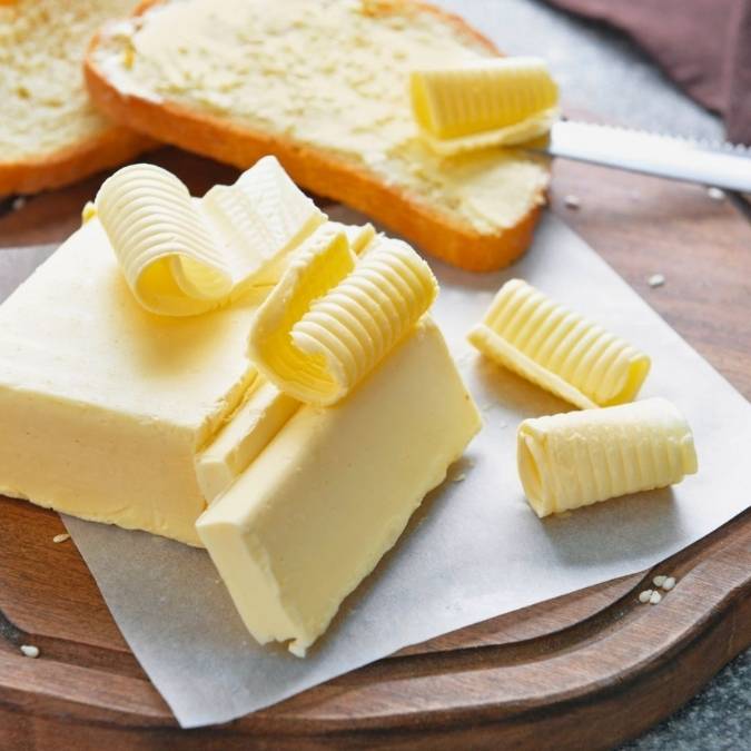 Blocks of pale yellow butter on parchment paper with curled butter decorations, alongside sliced toast topped with butter on a wooden board.