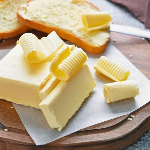 Blocks of pale yellow butter on parchment paper with curled butter decorations, alongside sliced toast topped with butter on a wooden board.