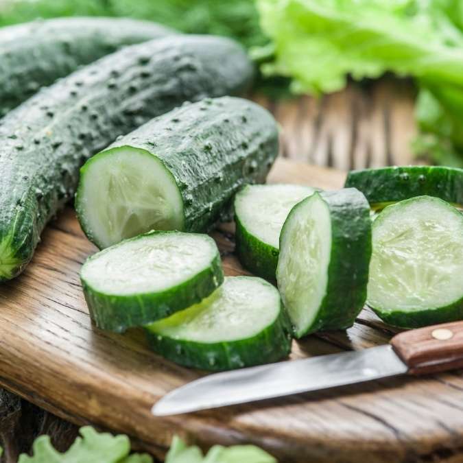 Fresh green cucumbers, including whole cucumbers and sliced rounds, arranged on a wooden cutting board with a small knife and leafy greens in the background.