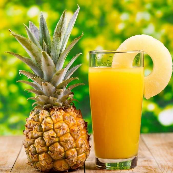 A whole pineapple sits next to a tall clear glass filled with bright orange pineapple juice, with a pineapple slice garnish on the rim, set on a wooden surface against a blurred green outdoor background.