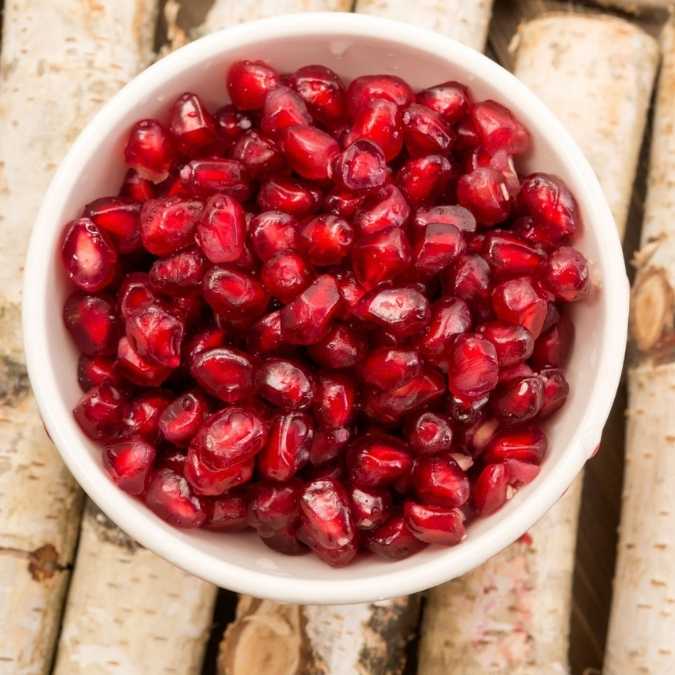 Glossy red pomegranate seeds filling a small white bowl, set against a rustic background of light-colored birch logs.