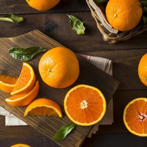 Fresh whole and sliced oranges arranged on a dark wooden cutting board and table, with a few green leaves scattered around. A woven basket holding whole oranges appears in the upper right corner.