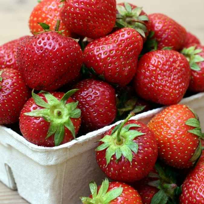 A close-up of fresh red strawberries piled in a white paper tray, with more strawberries visible around the edges.