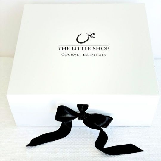 A white square gift box with the text 'THE LITTLE SHOP GOURMET ESSENTIALS' printed in black, tied with a black satin ribbon bow at the front, on a white textured surface.
