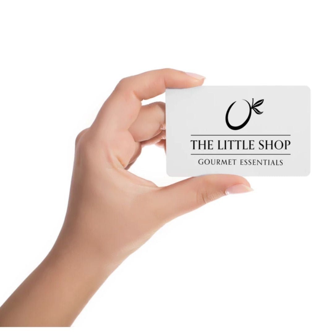 A hand holding a white gift card with the black printed logo and text 'THE LITTLE SHOP GOURMET ESSENTIALS' against a plain white background.
